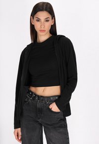 Crna zip-up dukserica preko crne kratke majice, uparena s visoko strukiranim sivim trapericama koje imaju pojas s okruglim detaljima.