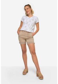 Camicia bianca a maniche corte con motivo geometrico beige, abbinata a pantaloni corti beige e sandali con zeppa color cuoio. Il modello si trova in piedi con le mani nelle tasche.