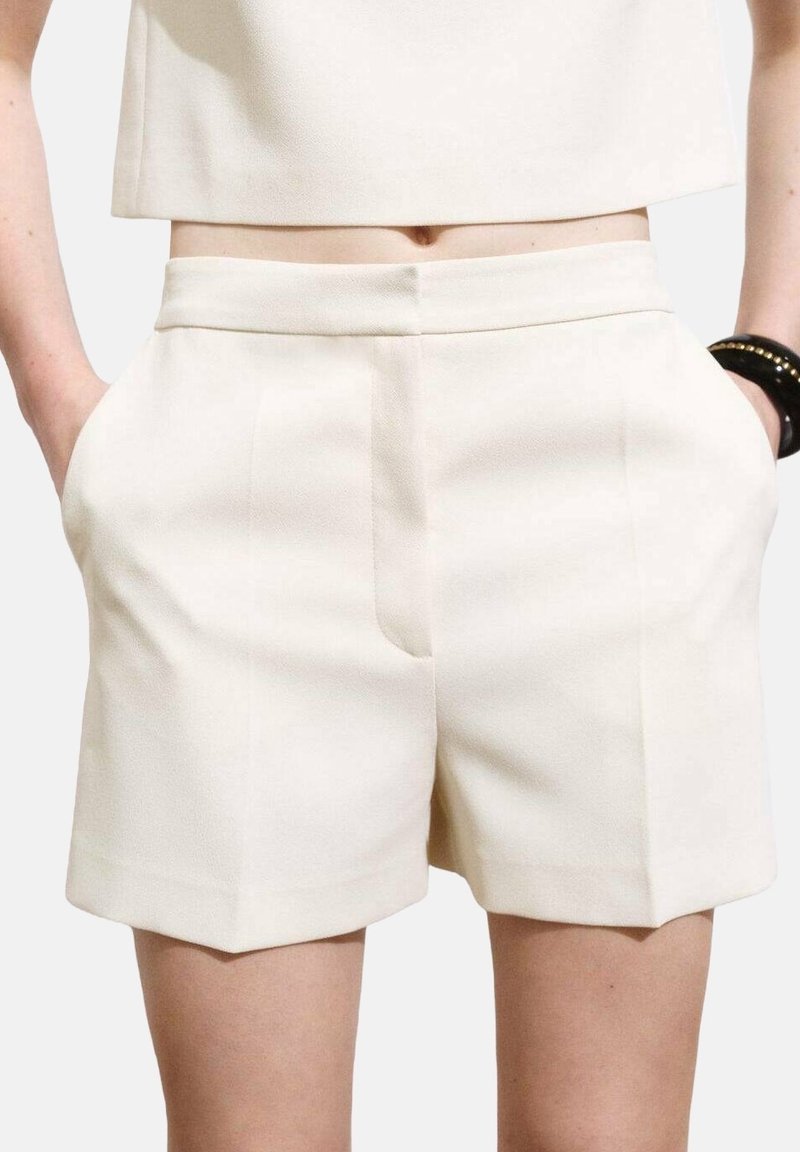 Femme portant un short blanc taille haute ajusté avec des poches, associé à un top court blanc et un bracelet noir au poignet.