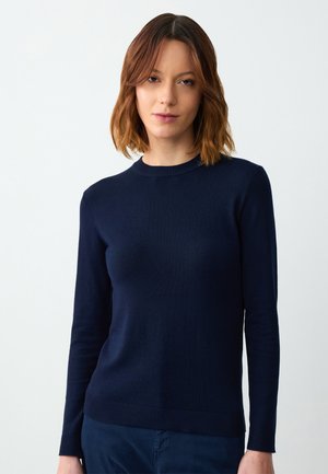 Donna con capelli castani lunghezza spalla che indossa un maglione fitted a maniche lunghe blu navy e pantaloni abbinati su uno sfondo semplice.