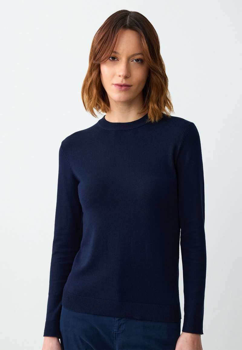 Kobieta z włosami do ramion w kolorze brązowym, ubrana w dopasowany granatowy sweter z długim rękawem i pasujące spodnie na jednolitym tle.