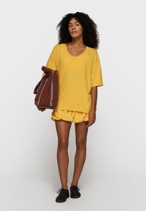 Kobieta o kręconych włosach, ubrana w żółtą, luźną koszulkę i szorty, trzyma dużą brązową torbę w paski, stoi na tle szarego tła.