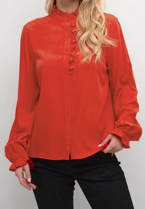 Femme portant une blouse rouge vif à manches longues avec col et poignets volantés, associée à un pantalon noir, sur un fond uni.