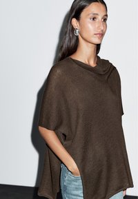 Kobieta z długimi ciemnymi włosami, ubrana w brązowy dziergany ponczo i srebrne owalne kolczyki, z jedną ręką w kieszeni jasnoniebieskich dżinsów, na tle białej ściany.