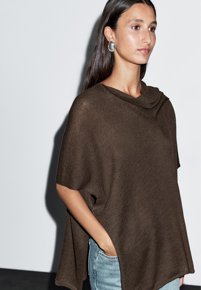 Kobieta z długimi ciemnymi włosami, ubrana w brązowy dziergany ponczo i srebrne owalne kolczyki, z jedną ręką w kieszeni jasnoniebieskich dżinsów, na tle białej ściany.