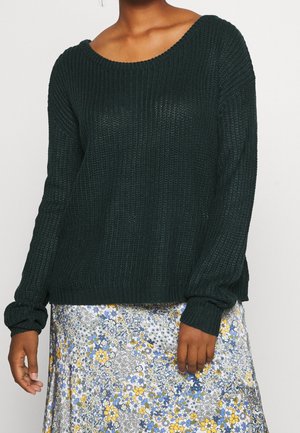 Personne portant un pull en maille vert foncé à manches longues et une jupe fleurie avec des fleurs jaunes, bleues et grises sur fond blanc.