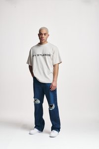 Smėlio spalvos medvilninis per didelis T-shirt su juodu tekstu "2Y STUDIOS", derinamas su mėlynomis džinsinėmis kelnėmis su pažeidimais ir baltais sportbačiais.