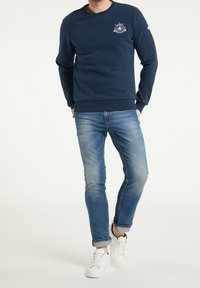 Granatowa bluza z okrągłym dekoltem i wyhaftowanym logo, zestawiona ze szczupłymi, jasnoniebieskimi dżinsami oraz białymi trampkami.