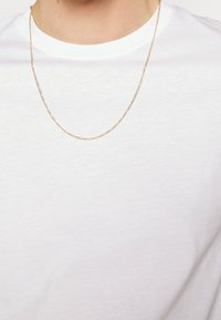 Collier en or avec un design délicat et entrelacé, porté sur un t-shirt blanc à col rond basique. Texture lisse et finition brillante.