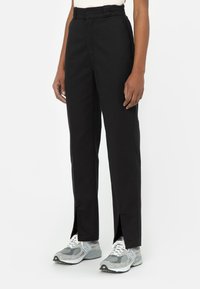 Pantalones negros de cintura alta con aberturas laterales en el dobladillo, hechos de una tela suave. Se llevan con zapatillas deportivas grises que tienen una suela blanca.