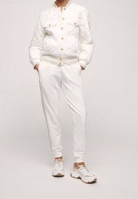 White quilted bomber jacket with gold buttons, paired with white joggers featuring gold stripes. Worn with white and tan sneakers.