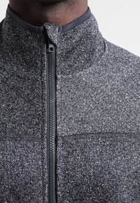 Giacca zip-up grigia scura con un motivo herringbone testurizzato, colletto alto e tessuto liscio a contrasto nella zona del collo e della cerniera.