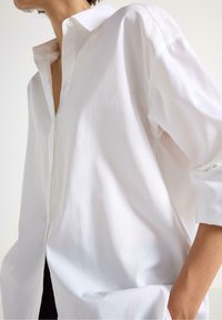 Person i en løs hvid skjorte med optrillede ærmer, hånd i lommen, mod en ensfarvet lys baggrund.