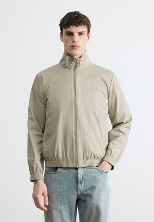 Νεαρός άνδρας που φοράει μπεζ μπουφάν με φερμουάρ, ελαστικά μανίκια και τελείωμα, και ανοιχτόχρωμα μπλε τζιν, στέκεται μπροστά σε ένα απλό λευκό φόντο.