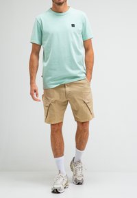 Light mint green short-sleeve t-shirt with a small black logo. Beige cargo shorts with two side pockets. White socks and chunky gray sneakers.
