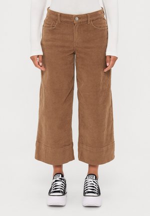 Pantalon large en velours marron porté avec des baskets Converse à plateforme noires et blanches et un haut blanc à manches longues, torse inférieur visible.