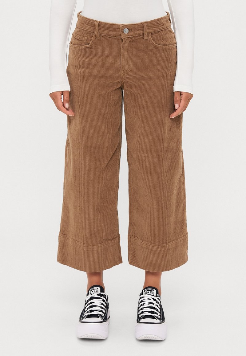 Pantalon large en velours marron porté avec des baskets Converse à plateforme noires et blanches et un haut blanc à manches longues, torse inférieur visible.