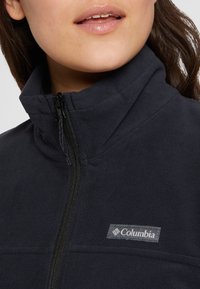Navy fleece takki, jossa on korkea kaulus ja etuvetoketju. Ominaisuuksiin kuuluu harmaa Columbia-logo rinnassa ja pehmeä, teksturoitu kangas.