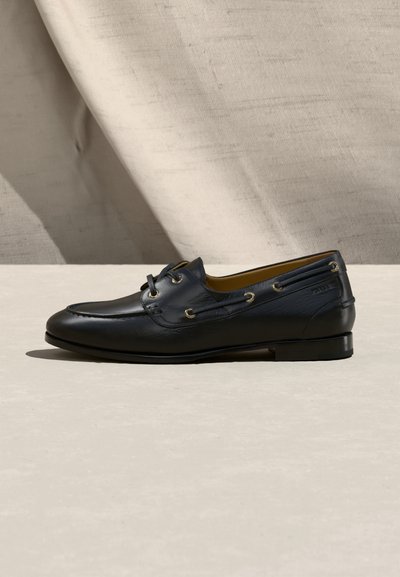 Chaussure bateau en cuir noir avec lacets latéraux, couture moc toe et petit talon, posée sur un fond de tissu neutre et une surface lisse.