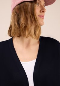 Navyfarbener Cardigan aus weichem Strickmaterial mit V-Ausschnitt, getragen über einem weißen Oberteil und ergänzt durch eine hellpinkfarbene Cordmütze.