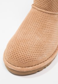 Beżowe zamszowe buty z perforowanym wierzchem i grubą podeszwą, przedstawione z bliska z przodu na białym tle.