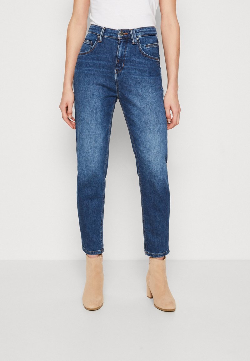 Jeans de denim azul de cintura alta con un acabado desgastado, cinco bolsillos y detalles de costura. Combinados con botines beige.