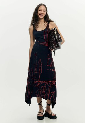 Junge Frau mit lockigem Haar, die ein schwarzes Kleid mit roten abstrakten Linien trägt, eine große schwarze Handtasche hält und schwarze Plateausandalen trägt.