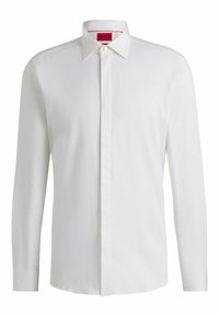 Camicia bianca a maniche lunghe con bottoni, dotata di un leggero lucido, colletto strutturato e pattina dei bottoni nascosta. Etichetta visibile di colore rosso all’interno del colletto.