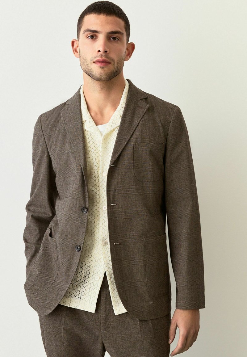 Braun karierter Blazer mit zwei Fronttaschen, getragen über einem cremefarbenen Hemd mit geometrischem Muster. Glatter Stoff mit taillierter Passform.