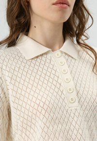 Gebreide polo in crème met een ruitpatroon, voorzien van een effen kraag en zes crèmekleurige knopen aan de voorkant. Lichtgewicht en met structuur.