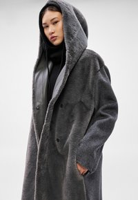 Šedý nadrozmerný kabát s kapucňou, s textúrovaným, plyšovým vonkajškom a hladkými koženými detailmi. Obsahuje bočné vrecká a dvojradové zapínanie.