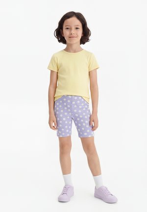 Niño de pie vistiendo una camiseta amarilla de manga corta, pantalones cortos morados con estampados de margaritas blancas, calcetines blancos y zapatillas deportivas moradas sobre un fondo blanco.