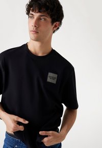 Juodas medvilninis T-shirt su kvadratiniu užkliju, kuriame užrašas "GUESS EST. 1981." Apskritas iškirptės ir trumpomis rankovėmis, pristatyta lengvame fone.