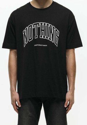 Osoba w czarnej koszulce z dużym białym napisem "NOTHING" oraz mniejszym napisem "GOODFORNOTHING" poniżej, stojąca z rozluźnionymi ramionami.