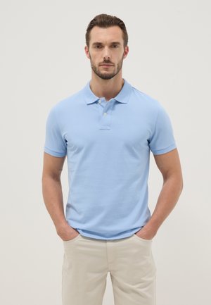 Homme avec barbe portant un polo bleu clair à manches courtes et un pantalon beige, debout les mains dans les poches devant un fond uni.