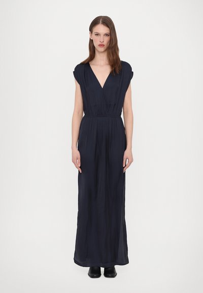 Maxi dress blu navy con scollatura a V, maniche a cappuccio, vita stretta e tessuto fluente. Indossato con stivaletti neri alla caviglia.