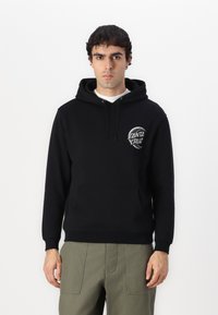 Felpa nera con cappuccio e tasca frontale, con un design circolare bianco del logo sul lato sinistro del petto. Il tessuto sembra morbido e spesso.