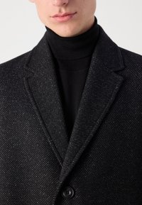 Cappotto in misto lana nera con un motivo a strisce diagonali, caratterizzato da un colletto a punta e una chiusura con un solo bottone. Tessuto testurizzato con una leggera lucentezza.