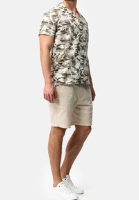 Kurzärmliges Hemd mit Knöpfen und grün-weißem abstraktem Blattmuster. Kombiniert mit hellbeigen Shorts und weißen Sneakers.