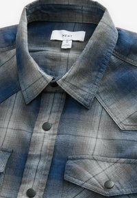 Niebiesko-szara koszula flanelowa w kratę z kołnierzykiem, czarnymi zatrzaskami i dwoma kieszeniami na piersi. Metka z napisem "NEXT", rozmiar M, wyprodukowano w Bangladeszu.