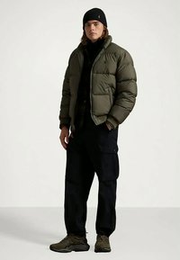 Nuori mies, joka on pukeutunut oliivinvihreään pakkasjakkuun, mustiin cargo-housuihin, mustaan pipoon ja tummiin lenkkikengiin, seisoo yksinkertaista valkoista taustaa vasten.