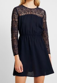 Marineblaues Kleid mit langen Spitzenschäften und tailliertem Schnitt. Verfügt über einen runden Ausschnitt und einen fließenden Rock, der oberhalb des Knies endet.