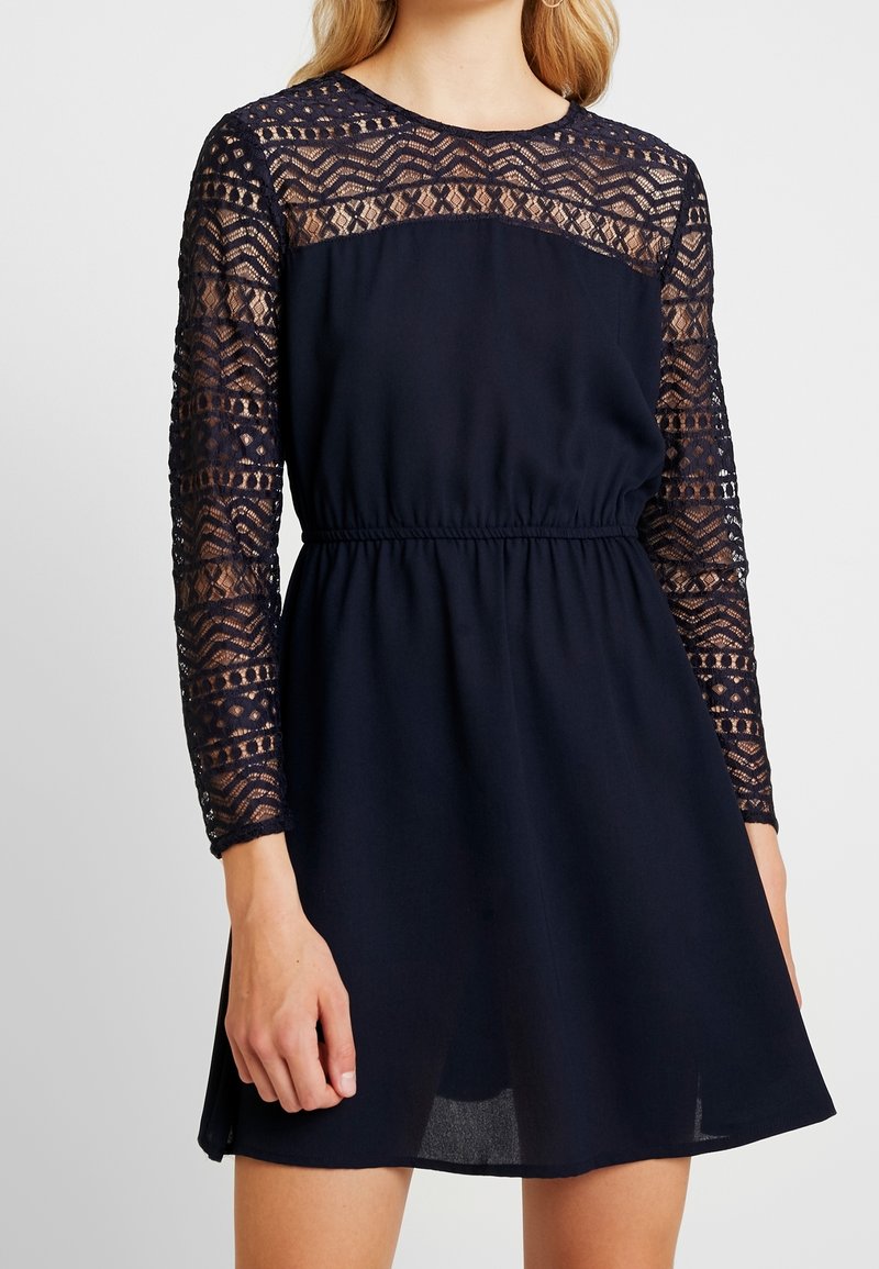 Marineblaues Kleid mit langen Spitzenschäften und tailliertem Schnitt. Verfügt über einen runden Ausschnitt und einen fließenden Rock, der oberhalb des Knies endet.