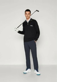 Pulover golf negru, cu fermoar la jumătate, având un guler alb și logo-ul "BOSS". Asortat cu pantaloni închiși la culoare și încălțăminte de golf albă.