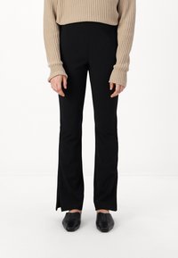 Pantalons noirs évasés avec une texture lisse, associés à un pull beige à côtes avec des manches longues et des poignets larges. Chaussures noires à bout carré.