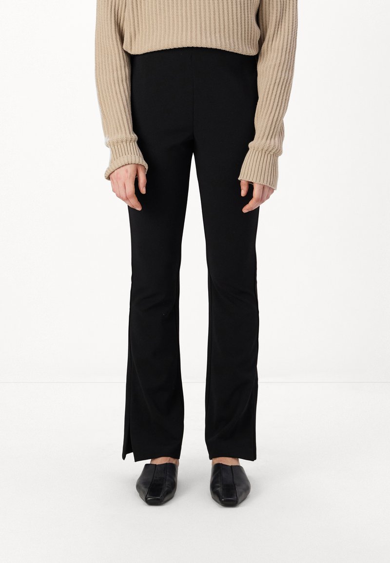 Pantalons noirs évasés avec une texture lisse, associés à un pull beige à côtes avec des manches longues et des poignets larges. Chaussures noires à bout carré.