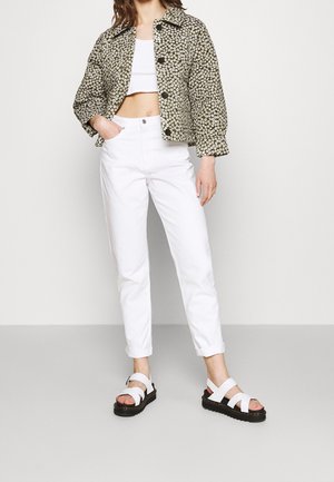 Vrouw draagt een korte bloemenjas over een wit cropped topje, witte hoge taille broek en witte sandalen met zwarte zolen.