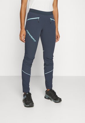 Person trägt marineblaue Sporthose mit hellblauen Reißverschlüssen und Logo, kombiniert mit schwarzen und grauen Wanderschuhen vor weißem Hintergrund.