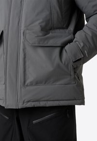 Grå isolerad jacka med ett lätt tyg, stora framfickor, justerbara ärmslut och en vattentålig ytbehandling.