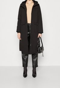 Manteau long matelassé noir avec capuche, fermeture éclair à l'avant et poches ; porté sur un haut beige et un pantalon en simili cuir noir. Bottines à talons noires et sac à main.